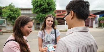 A photo of a pair of sister missionaries in Thailand speak to a man on the street and handing him a passalong card